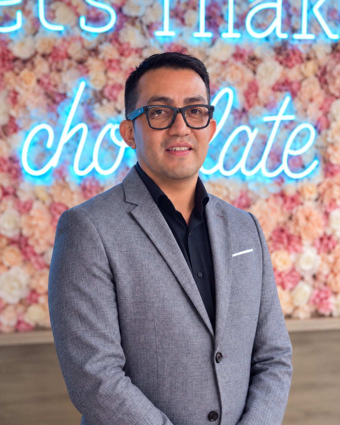 Jorge E. Molina, General Manager of The Conche, in front of a flower wall with a neon chocolate sign